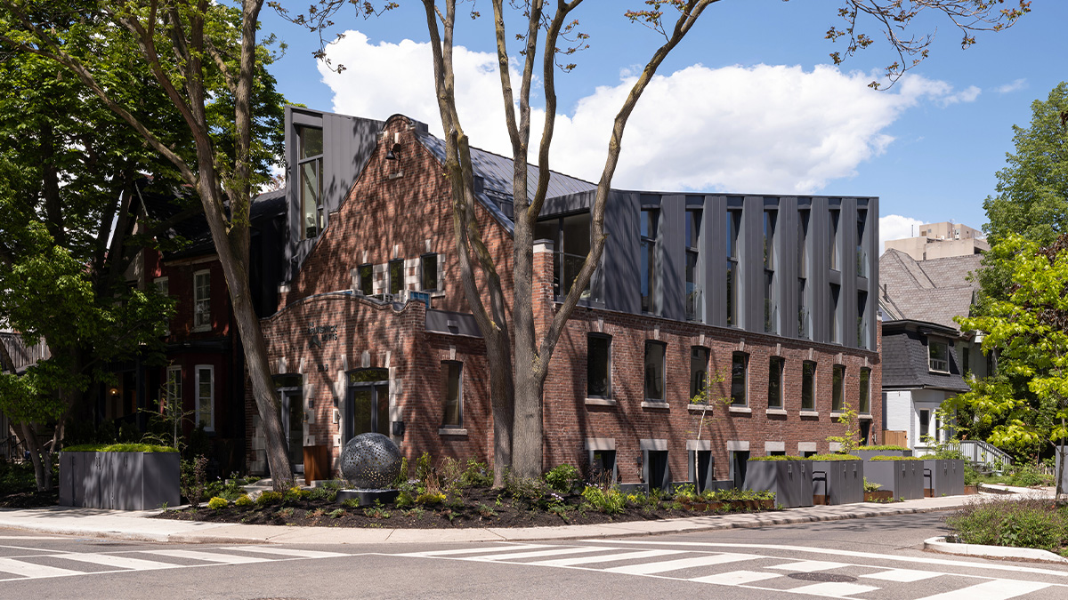 A modern brick building with large windows and metal cladding, surrounded by tall trees on a residential street corner