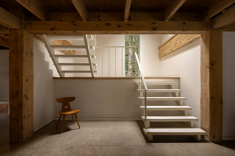 An interior space with exposed wooden beams, a white modern staircase leading to an upper level, and a wooden chair placed against the wall