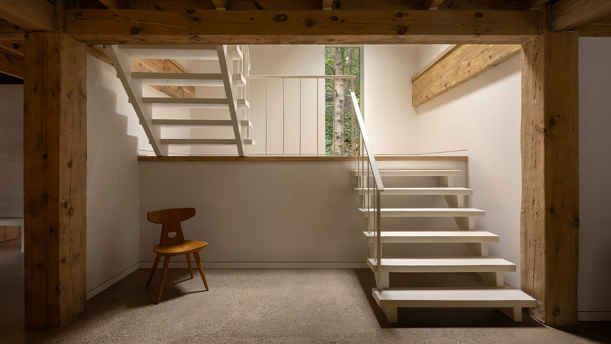 An interior space with exposed wooden beams, a white modern staircase leading to an upper level, and a wooden chair placed against the wall