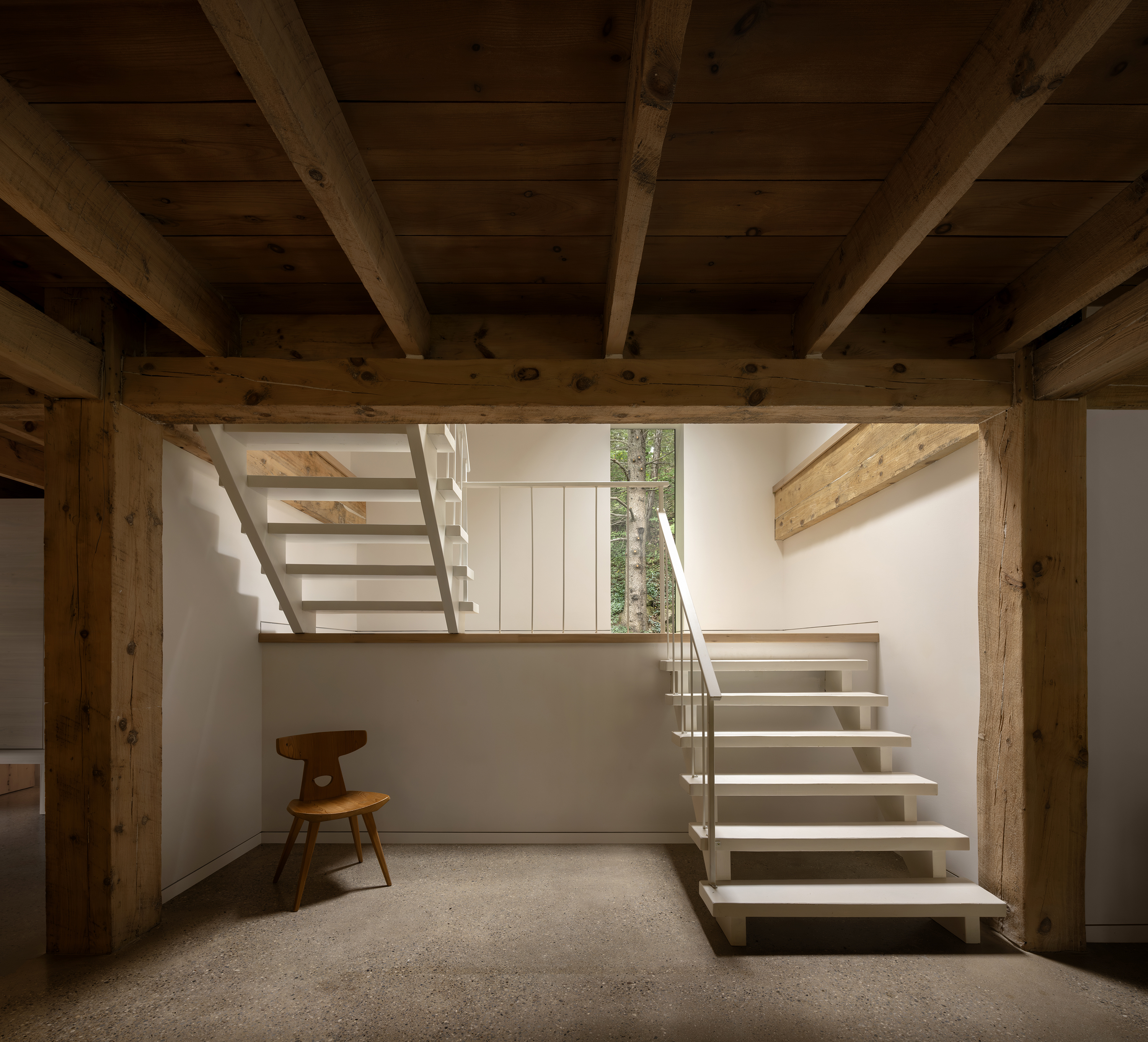 An interior space with exposed wooden beams, a white modern staircase leading to an upper level, and a wooden chair placed against the wall
