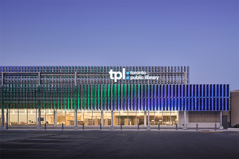 A public library building with a horizontal façade made of vertical metal fins in gradients of green, blue, and purple, large windows on the ground floor reveal bookshelves and interior lighting