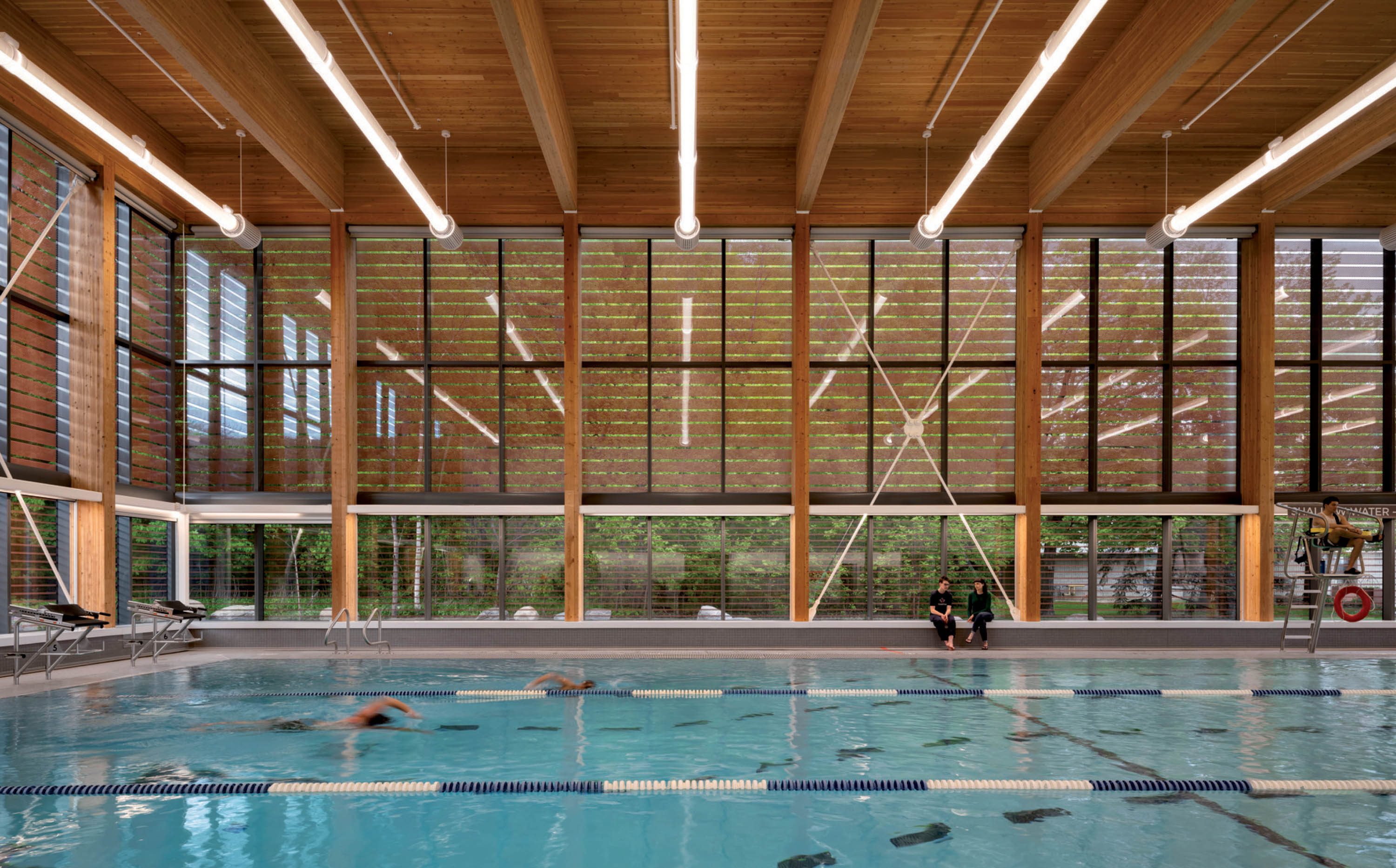 An indoor swimming pool with lane markings, surrounded by tall glass windows and exposed wooden beam