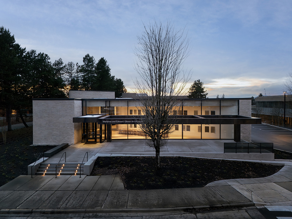 A modern art museum with a glowing entrance sits behind a paved walkway and sparse landscaping