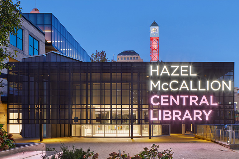 An illuminated modern library entrance at dusk featuring a dark metal façade with large glass panels and bright neon signage reading “Hazel McCallion Central Library,” with a clock tower and surrounding buildings visible in the background