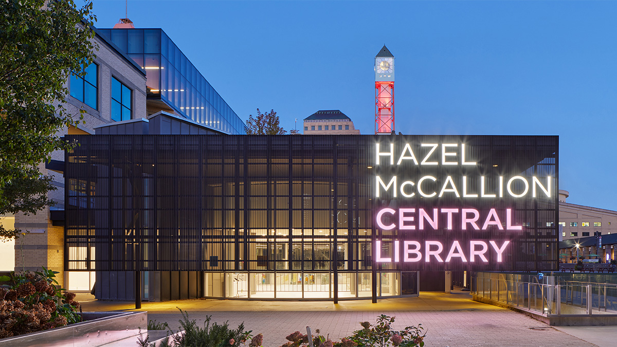 An illuminated modern library entrance at dusk featuring a dark metal façade with large glass panels and bright neon signage reading “Hazel McCallion Central Library,” with a clock tower and surrounding buildings visible in the background