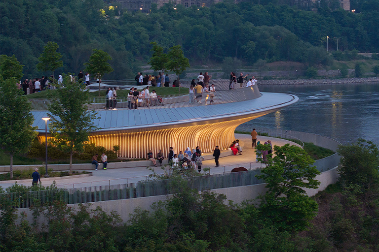 A curved, elevated waterfront overlook with a glowing underside, surrounded by trees and walking paths