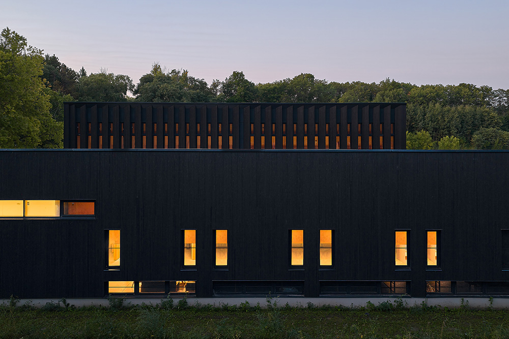 A dark, modern building glows warmly from tall rectangular windows at dusk