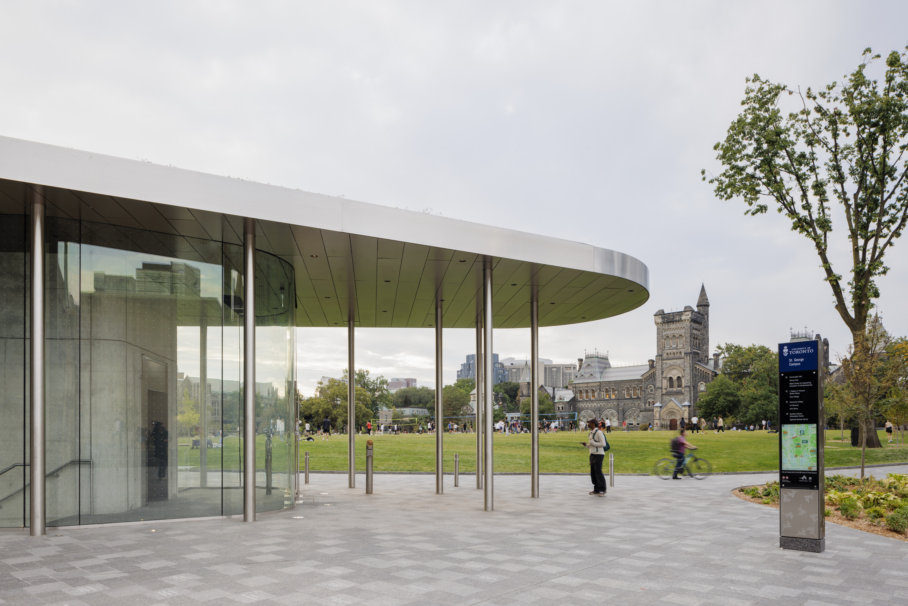 A paved public plaza with people walking near a sleek glass pavilion, with trees showing fall colors