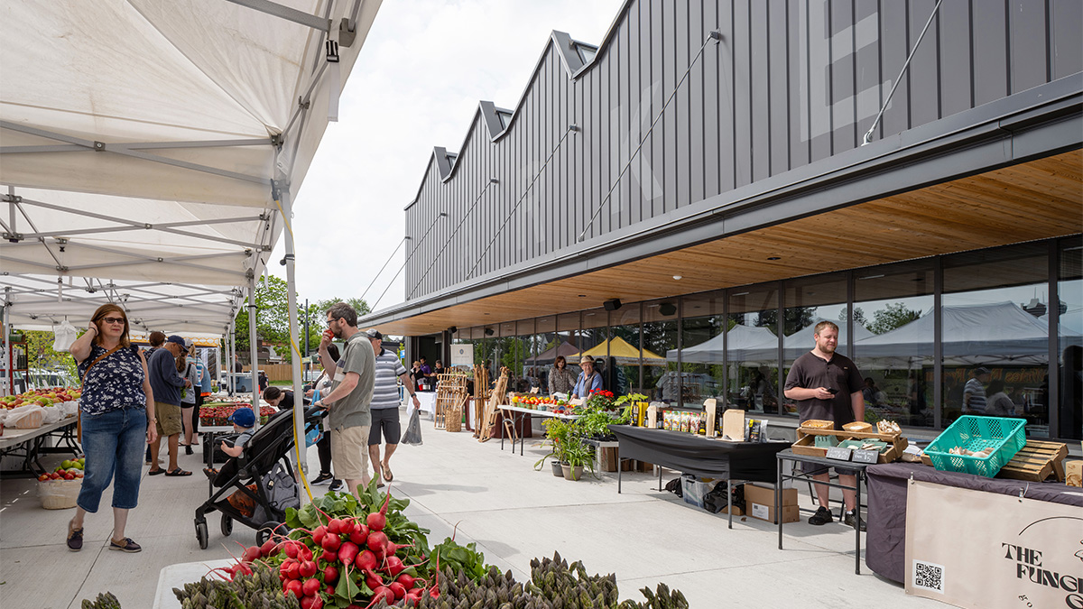 An outdoor farmers’ market with vendor tents set up along a building with a sawtooth roofline