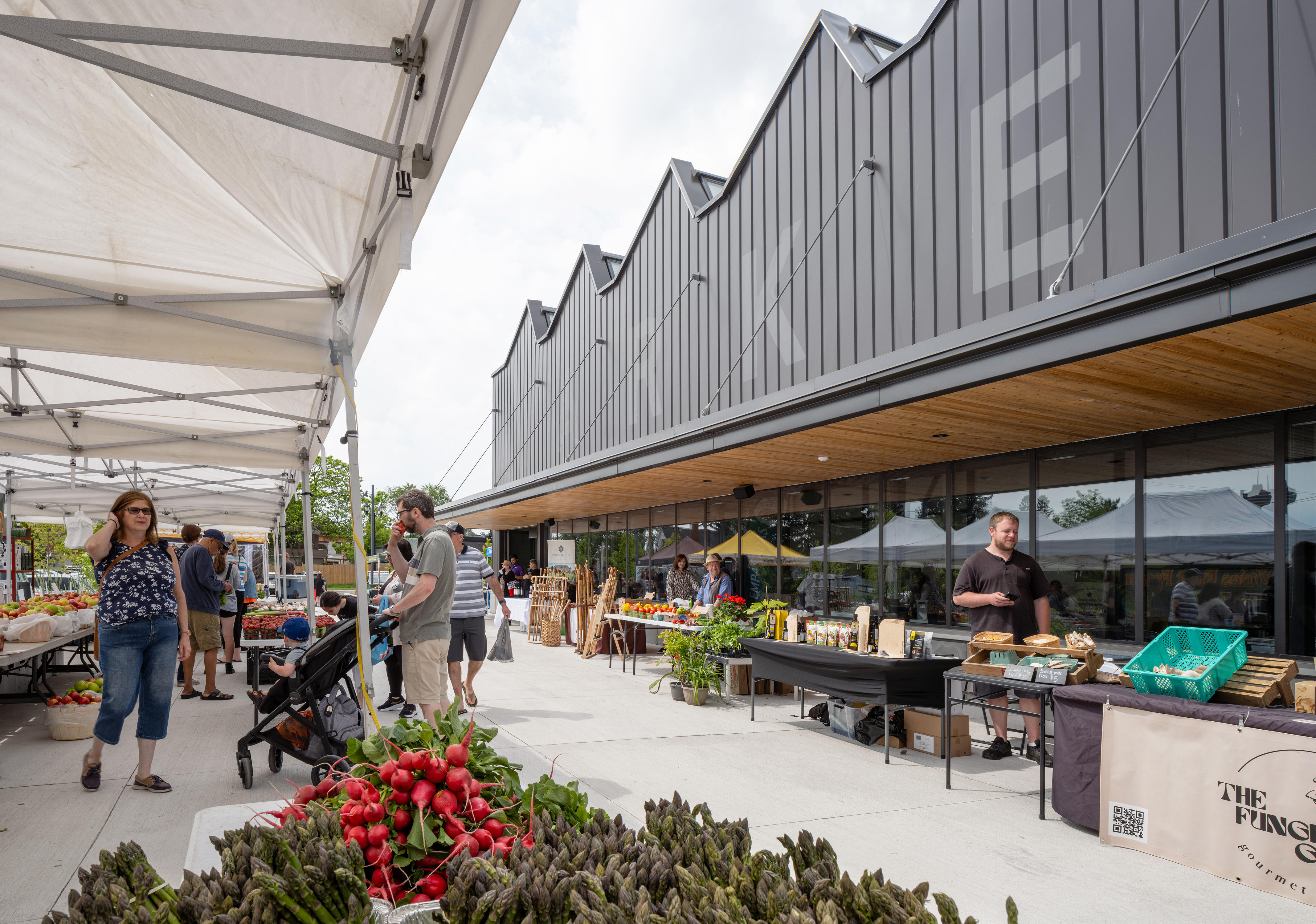 An outdoor farmers’ market with vendor tents set up along a walkway beside a building with a sawtooth roofline