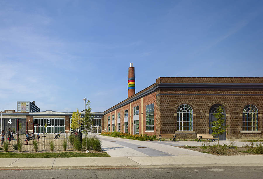 Kids play on a playground in front of five side-by-side red-brick warehouses.