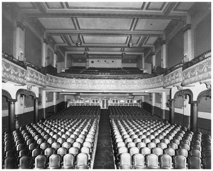 In one-point perspective and black and white, rows of seating extend to the back of a long narrow room with an aisle in the middle and columns lining the walls. A balcony at the back of the room is lined with decorative millwork and supports more chairs.