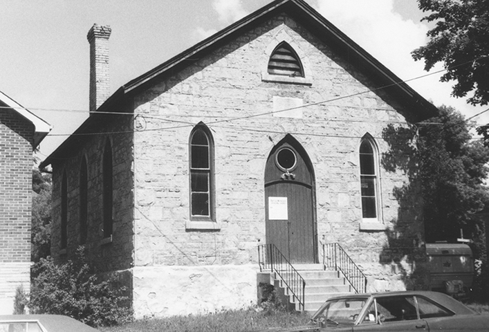 A limestone church with a gabled roof, a gothic gable vent, and two lancet windows on either side of a pointed arch front door reached by a set of stairs.