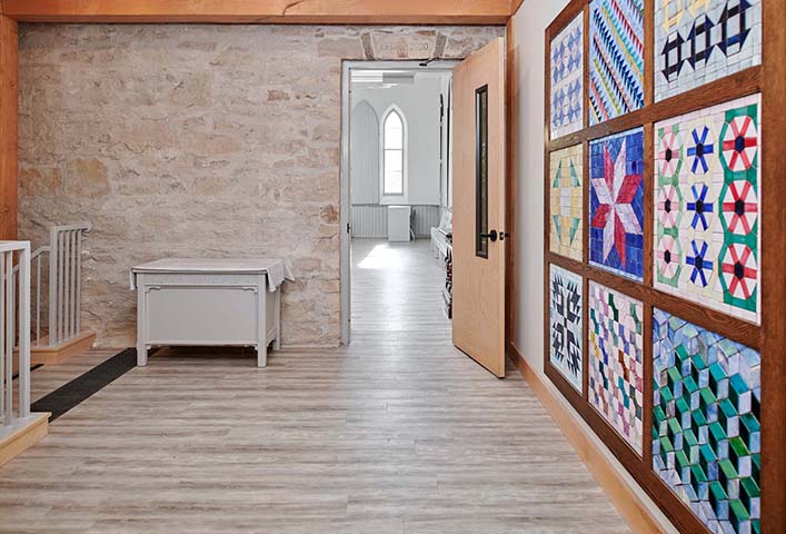 A one-point perspective in a brightly lit interior space with light wood flooring. Another brightly lit space is visible through an open wooden door in a stone wall and, on the right, a large grid of nine quilts is framed on the wall.