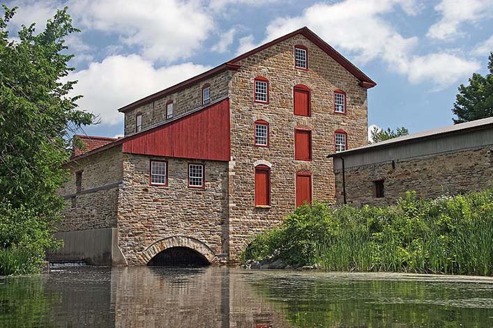 A stone building with red-painted wooden details stands over a river surrounded by greenery.