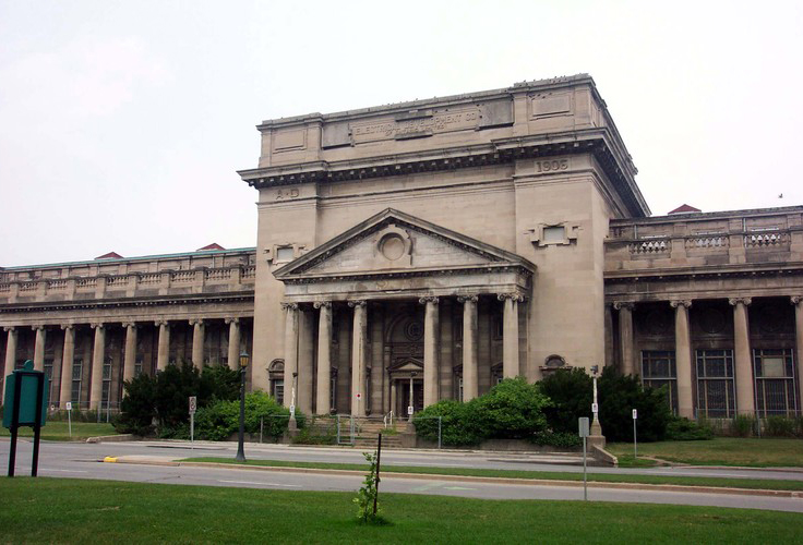 : A beige limestone stone building lines a street in front of a field. The ionic portico is centred in the frame as two ionic colonnades extend on both sides.