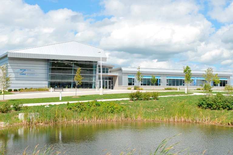 Exterior view of Walkerton Clean Water Centre’s building, a low one storey contemporary building with a large windows situated in front of a stormwater retention pond and young vegetation