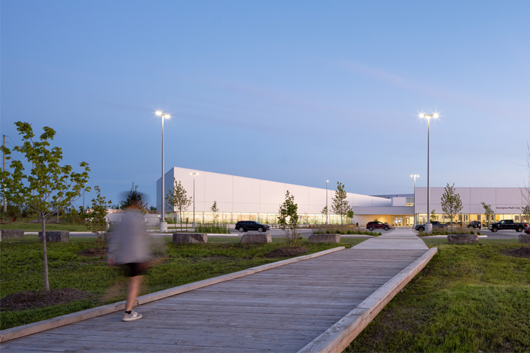 A wooden path overtop lush green grass leads to the modern looking Georgina Multi-Use Recreation Centre