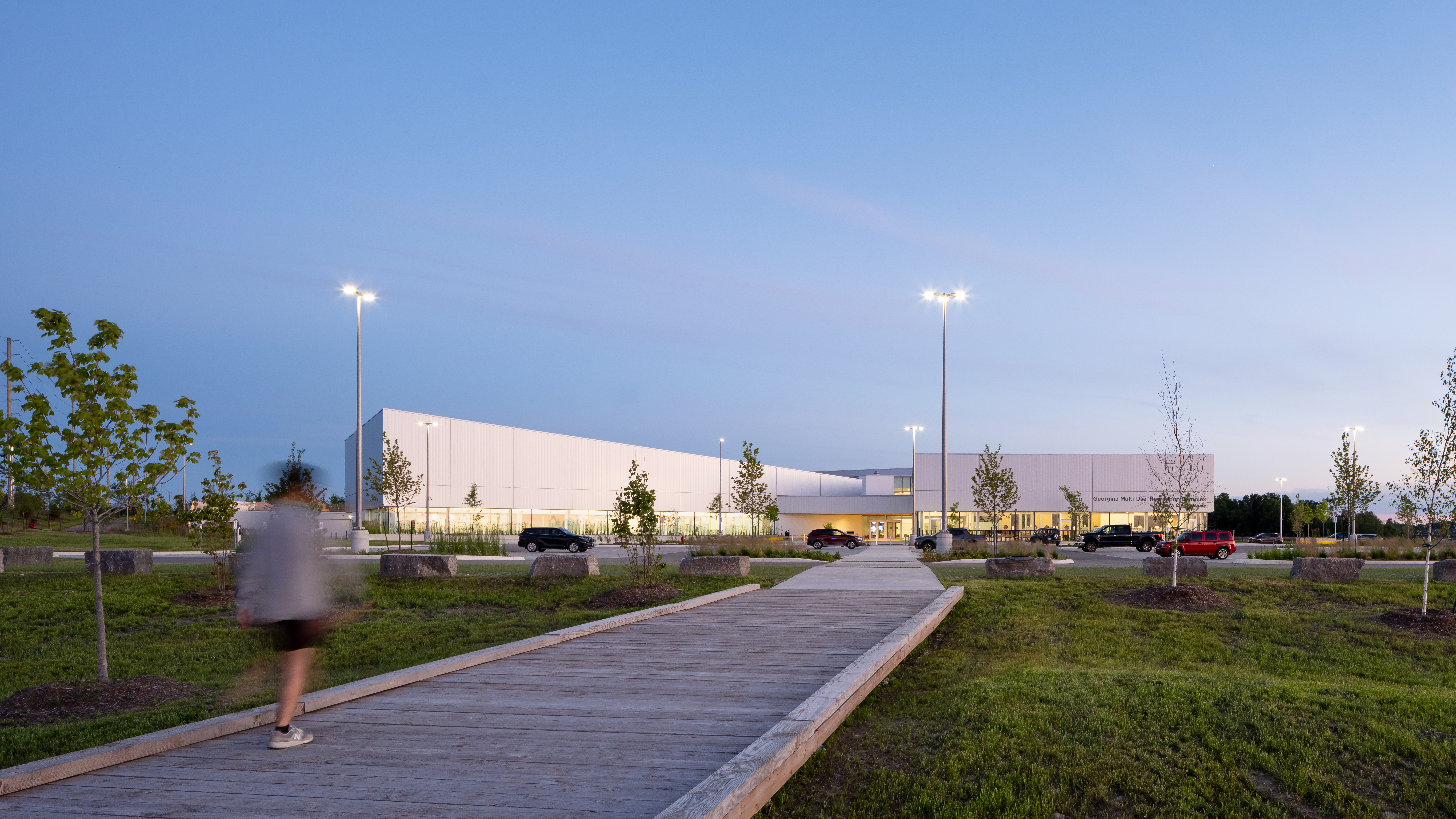 A wooden path overtop lush green grass leads to the modern looking Georgina Multi-Use Recreation Centre