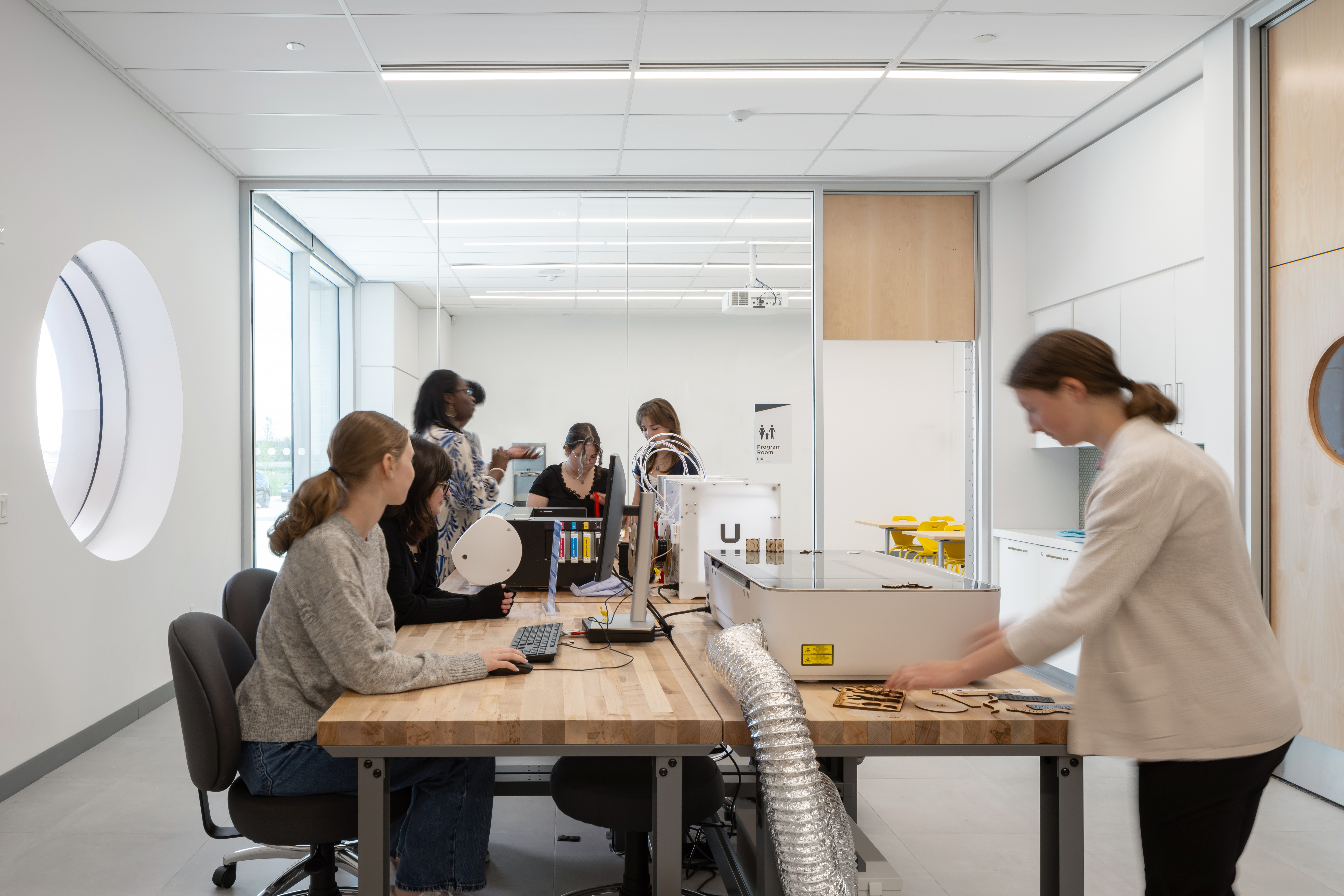 Bright workshop with a large table, computers, and printer. People collaborate on projects.
