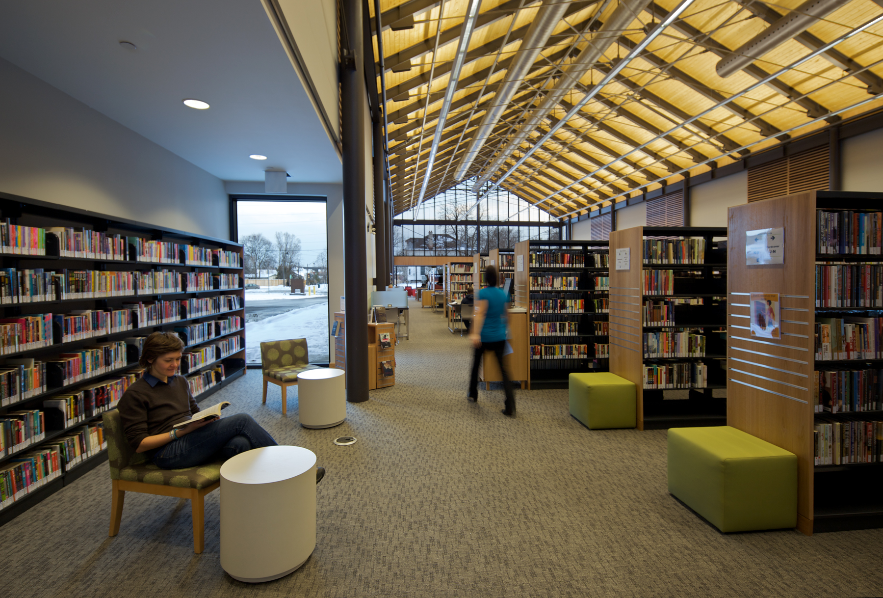 A person reading a book next to library stacks under an uplit wood and steel cathedral ceiling, resembling the inside of a barn.