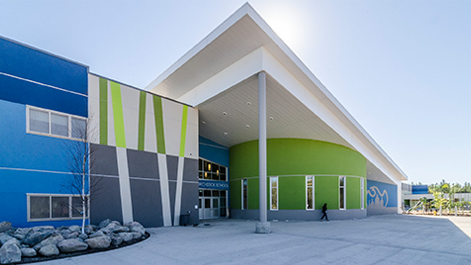 Large two-storey blue, grey, and green stucco building with a large, angled canopy stretching over the entrance.