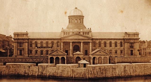 Historic sepia image of Kingston City Hall with heavy stone battery in front at a lower elevation. The central dome does not contain a clock.