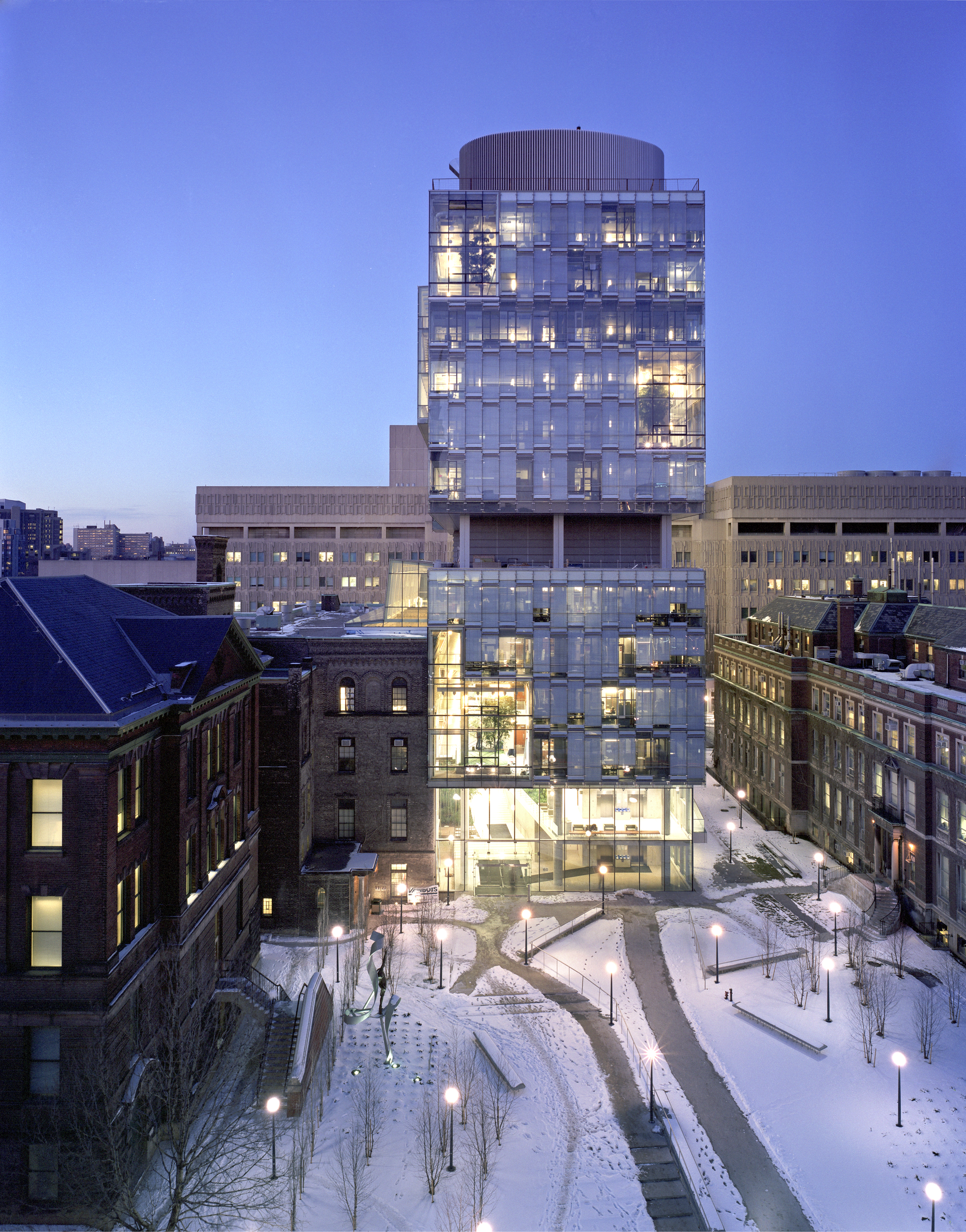 Front elevation of a tall, 12-storey building composed of two stacked glass boxes with a rounded metal mechanical penthouse on top. In front of the building, a snowy courtyard with pathways leading from the street to the building.													