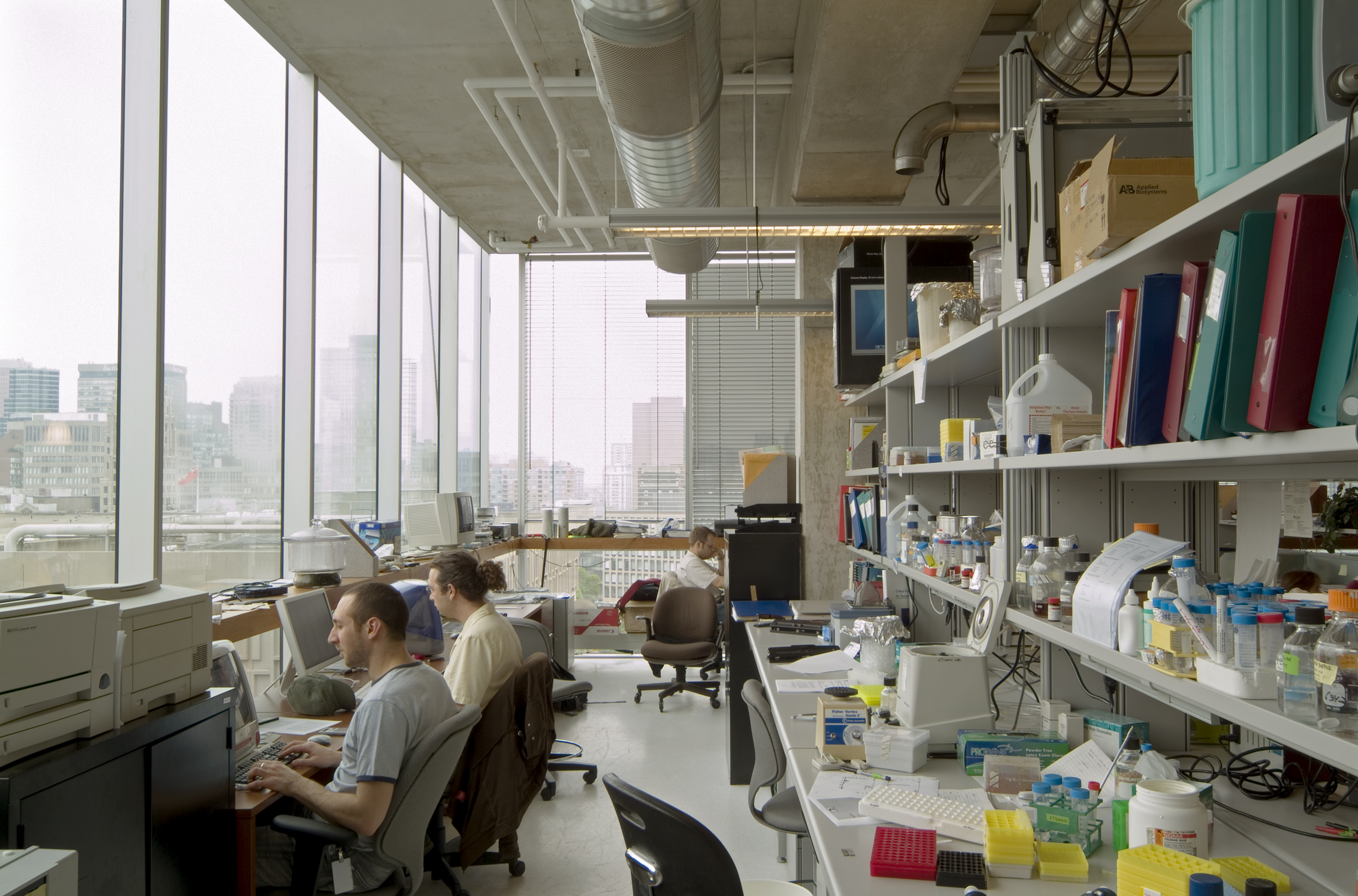 Three researchers sitting at at desks in front of full height curtain wall windows that wrap around a corner. Behind them are tables and shelves full of lab equipment and books.										