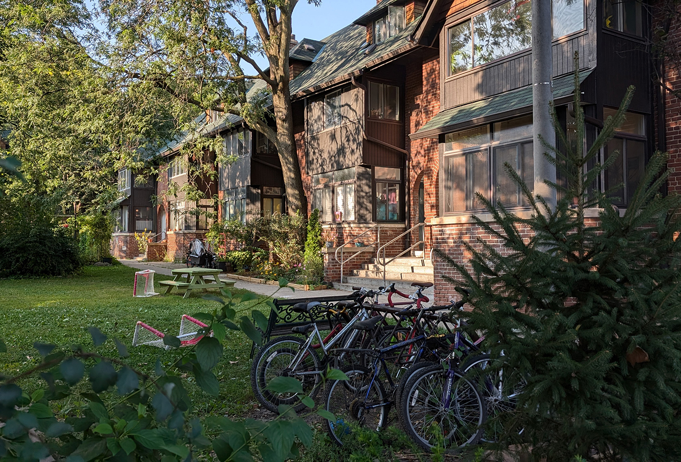  View of a row of houses in a green, shaded courtyard with large trees. The buildings have a mix of dark wood and brick facades with sloped roofs. Bicycles are parked in the foreground near a small spruce tree, and a picnic table and children’s mini hockey nets are set up on the lawn.