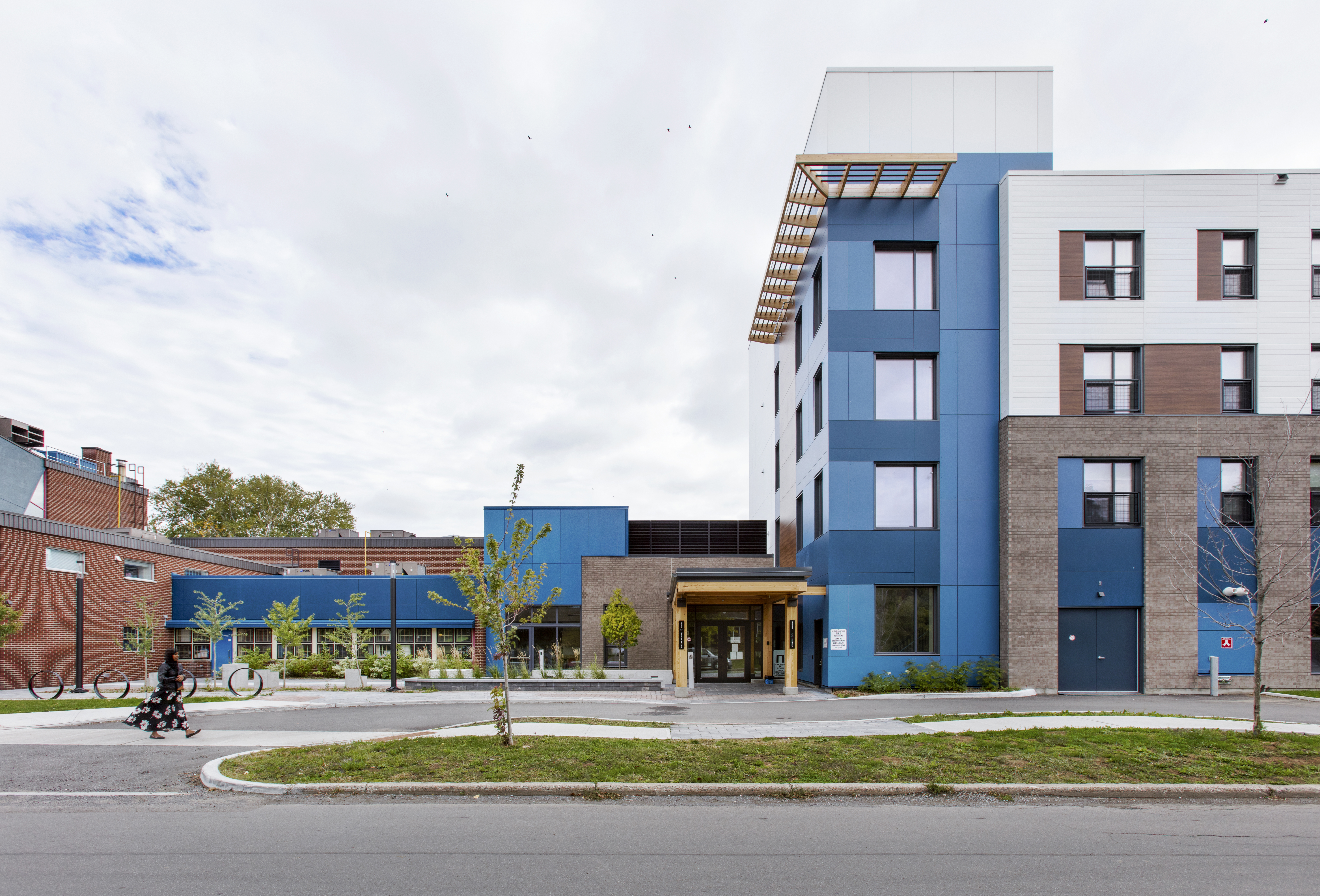 Street view of a modern mid-rise building with a mix of blue, white, and gray facades. The building features large windows and a wooden canopy over the entrance. The lower level has a brick exterior with a planted area in front. Young trees are planted along the street in front of the building.