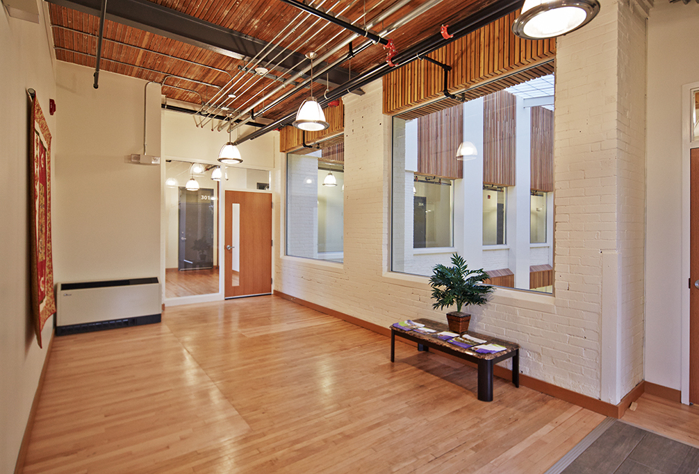 Interior view of a hallway featuring light wood floors, white brick walls, and large windows looking into an atrium. Exposed piping and industrial-style lighting hang from the wood-paneled ceiling. A small bench with brochures and a potted plant are positioned near the windows, creating a welcoming atmosphere.