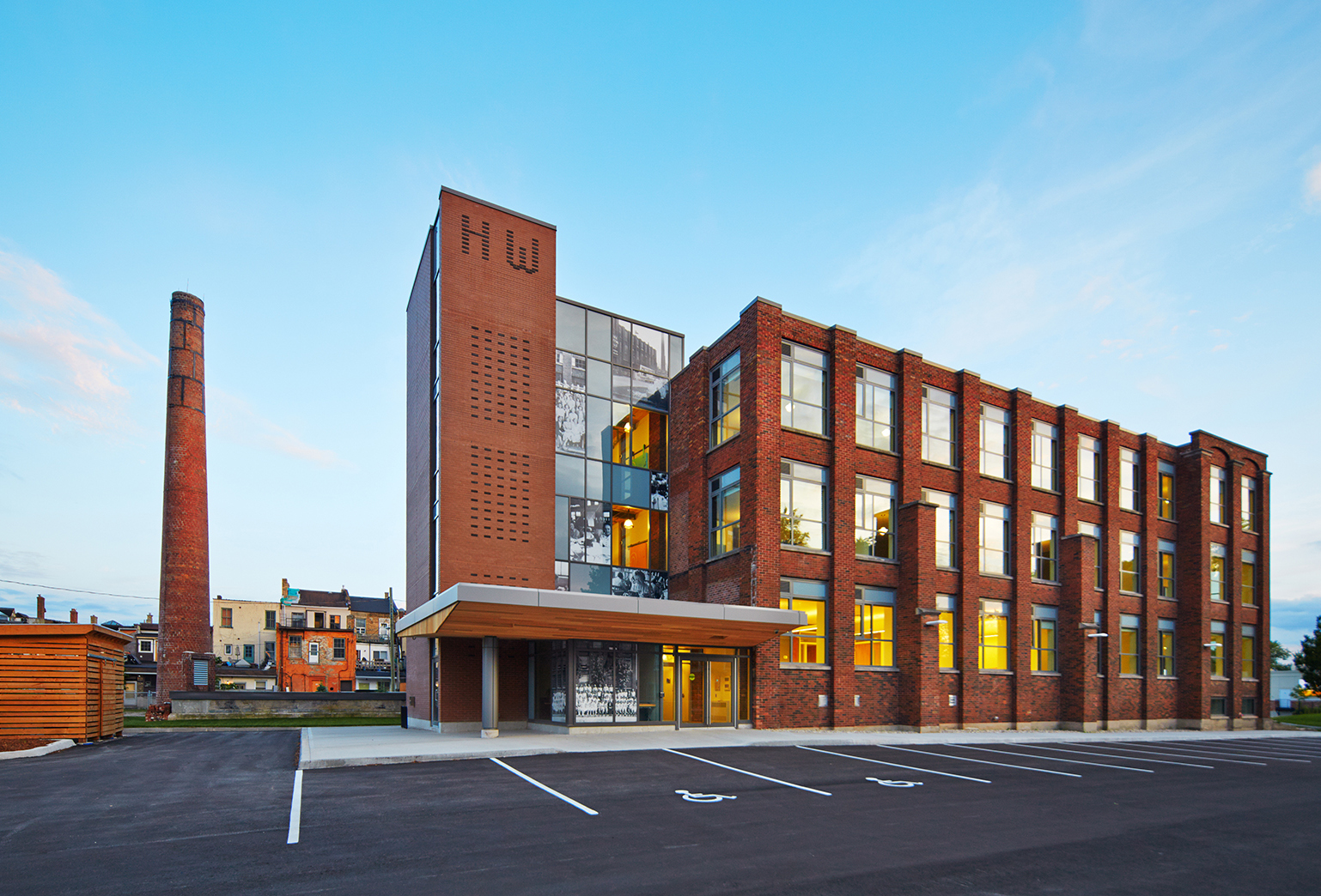 Exterior view of a renovated brick building with large windows and a modern entrance canopy. The building features a vertical brick tower with dark bricks forming the initials ‘HW’ and a glass section with historical photos. A tall, weathered brick smokestack is visible to the left, hinting at the building’s industrial past.