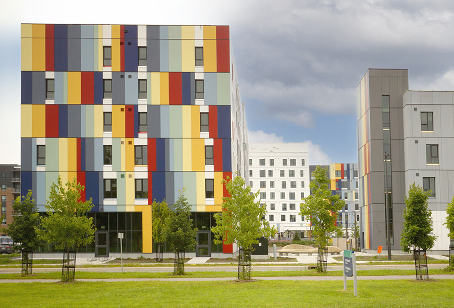 Wide-angle view of modern multi-story buildings with colorful facades featuring vertical stripes in shades of red, yellow, orange, blue, and gray. The buildings are part of a residential complex with large windows and green spaces in the foreground, including newly planted trees.