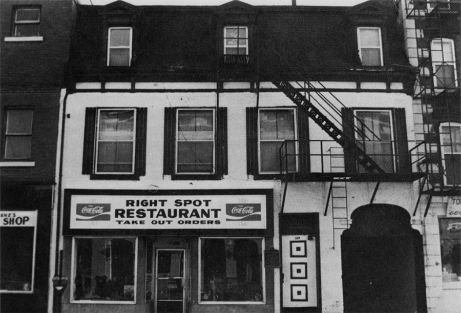 Black and white photo of a two-story building with a restaurant on the ground floor. The sign above the entrance reads ‘Right Spot Restaurant’ with a Coca-Cola logo. The facade features two large display windows and a central door. The upper floor has three windows with dark shutters, and a fire escape is visible on the right side of the building.