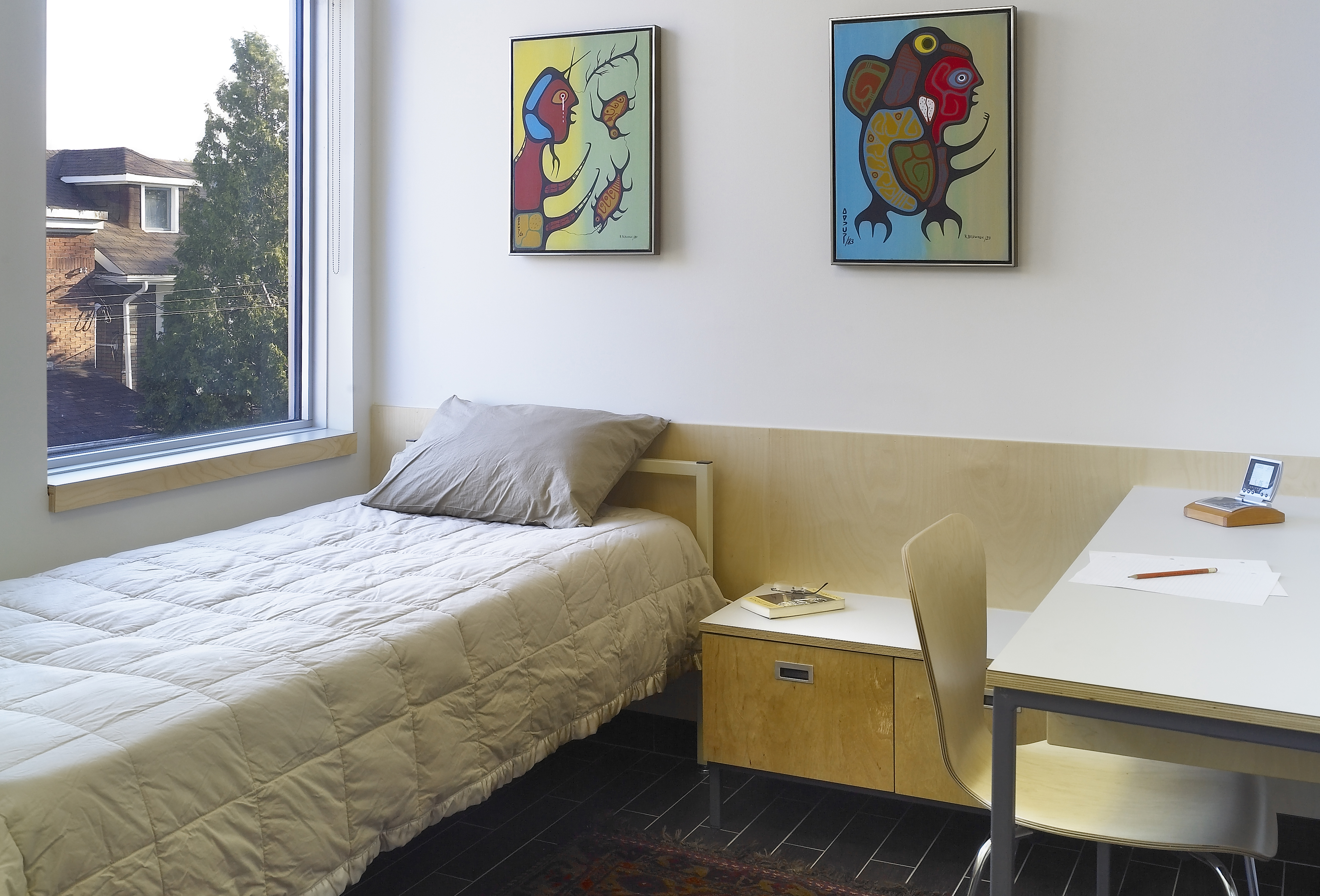 Interior of a simple, modern bedroom with a large window providing natural light. The room features a single bed, a small wooden nightstand, and a desk with a chair. Two colorful paintings hang on the wall above the bed. The floor is dark with a patterned rug.