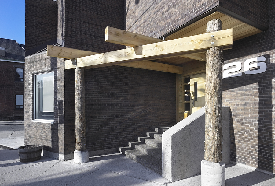 Entrance of a brick building with a distinctive wooden pergola supported by two rough-hewn wooden posts. The entrance features concrete steps leading to a recessed door, and the building number ‘26’ is displayed prominently on the brick wall beside the steps.