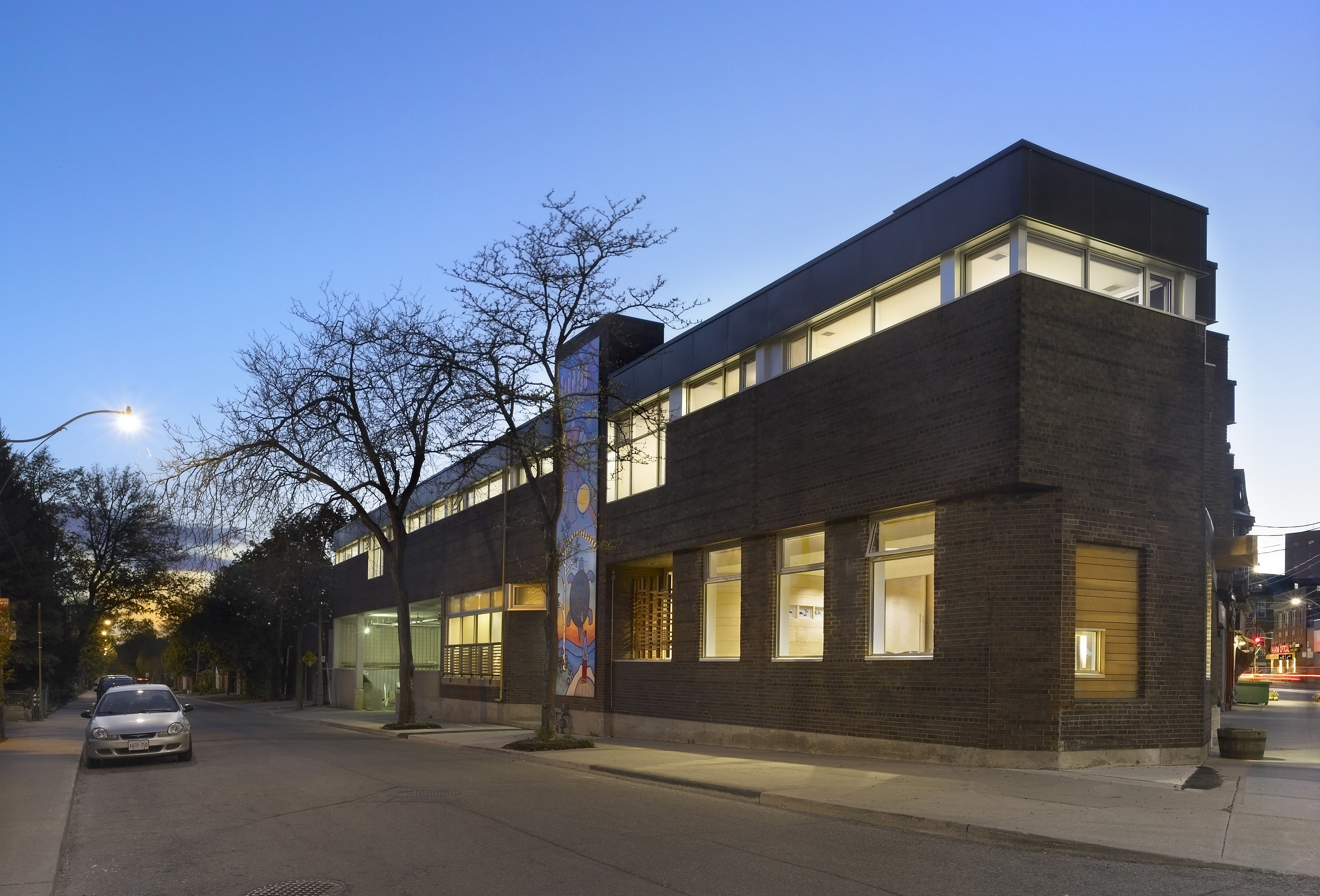 Exterior view of a brick two-story building with dark brick walls and large windows, taken during twilight. The upper windows wrap around the building and a vibrant mural is painted on the side of the building.