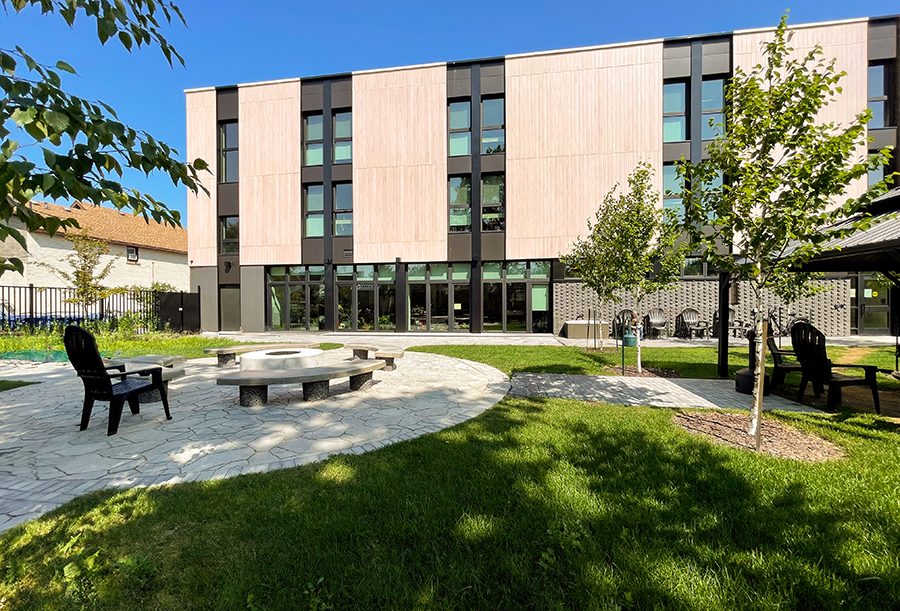 Exterior view of a modern three-story building with a light wood and black panel facade. In the foreground is a landscaped courtyard with grass, young trees, and a circular stone seating area surrounding a fire pit. Black outdoor chairs are positioned around the space, and large windows on the ground floor provide a view into the interior.