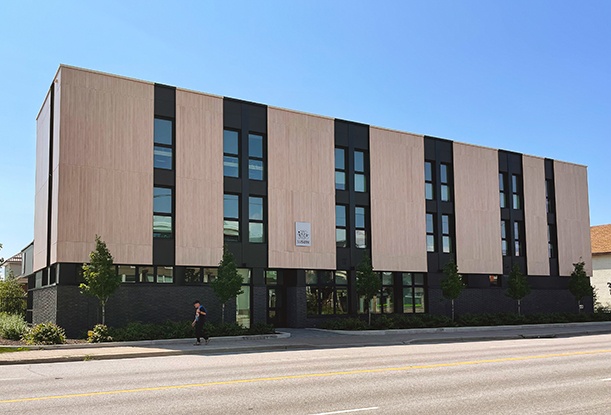 Street view of a modern three-story building with a light wood and black panel facade. The building has large vertical windows and a minimalist design. A sign with the name ‘Suswin’ is mounted on the facade. The lower portion of the building is clad in dark brick, with landscaping and young trees along the sidewalk.