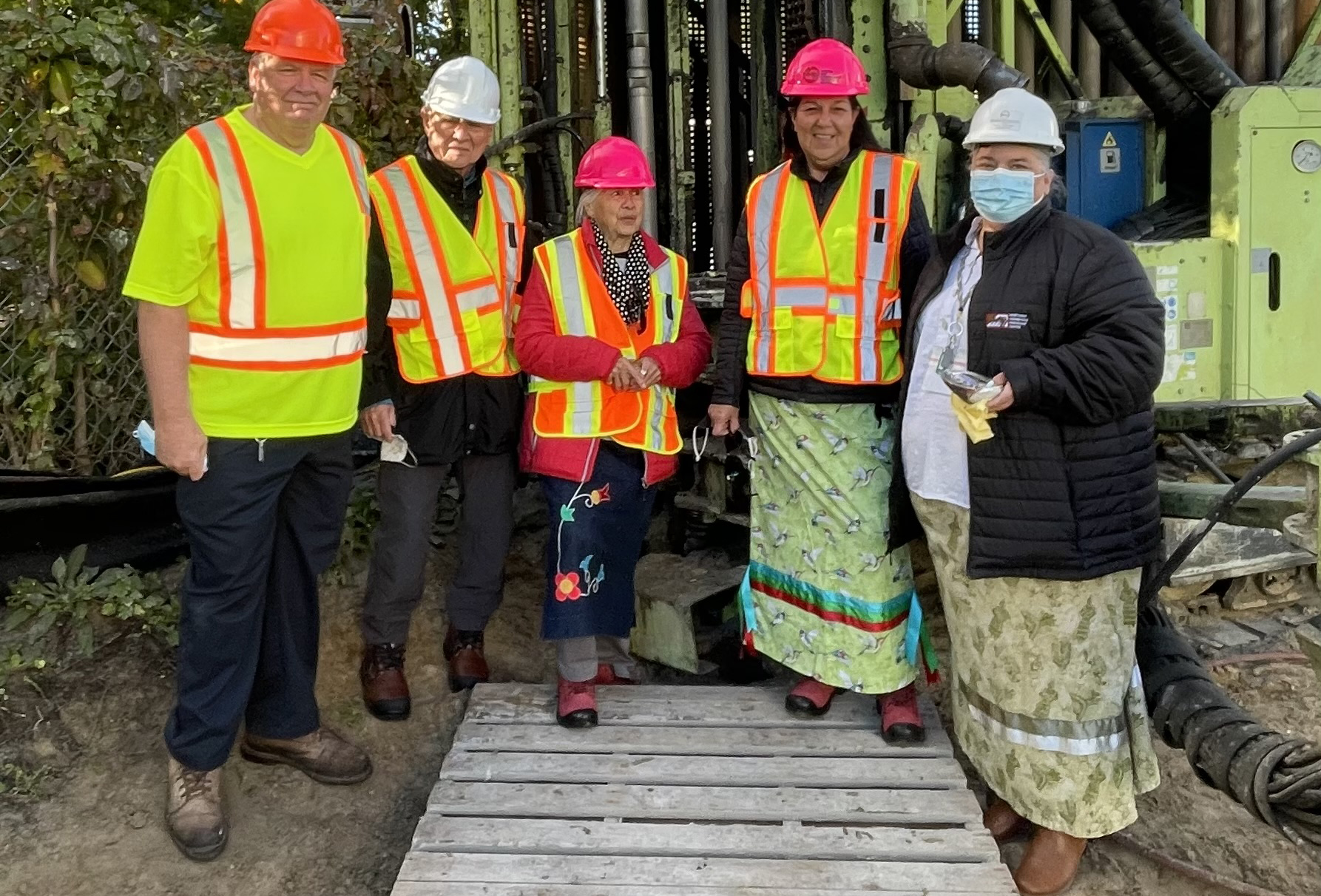 Group of five people wearing hard hats, high-visibility safety vests, and skirts standing in front of a large piece of industrial equipment at a construction site. The group is standing on a wooden platform, and trees are visible in the background.