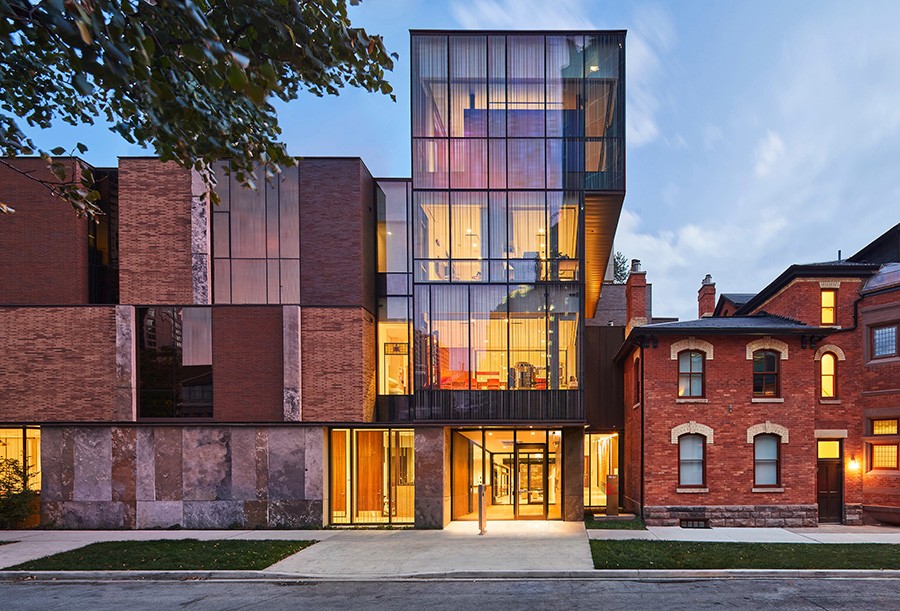 Exterior view of a modern building with large glass windows on the left and a restored historic red brick house on the right. The modern section has clean lines and a mix of brick, glass, and stone materials, with warm interior lighting visible through the windows. The historic house features traditional architectural details  such as arched windows and decorative brickwork.