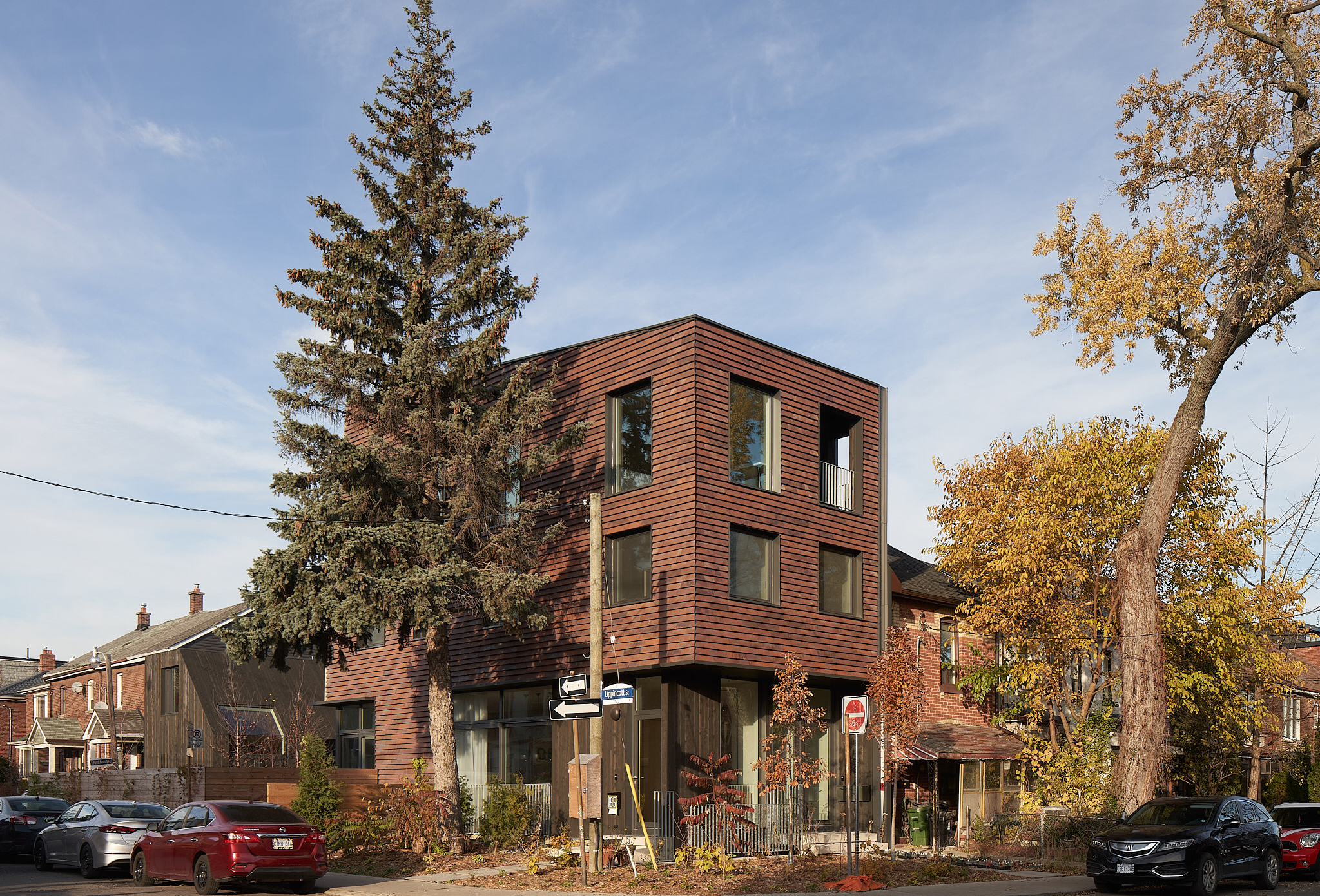 Exterior view of a modern, three-story corner house clad in horizontal clay tile. The building features large square windows and blends into the surrounding residential neighbourhood with mature trees. A tall evergreen tree stands in front of the house.