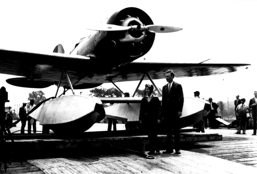 Black and white photo of a vintage seaplane with large pontoons, viewed from the front. Two people, one in aviator gear and the other in a suit, stand next to the plane. Other people are also visible in the background, gathered around the aircraft on a wooden dock.