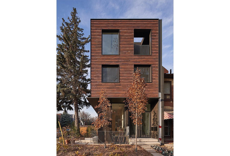 Front view of a modern, three-storey house with horizontal clay tile and large square windows. The building has a small recessed balcony. The ground floor features floor-to-ceiling windows. A large evergreen tree and young deciduous trees frame the house.