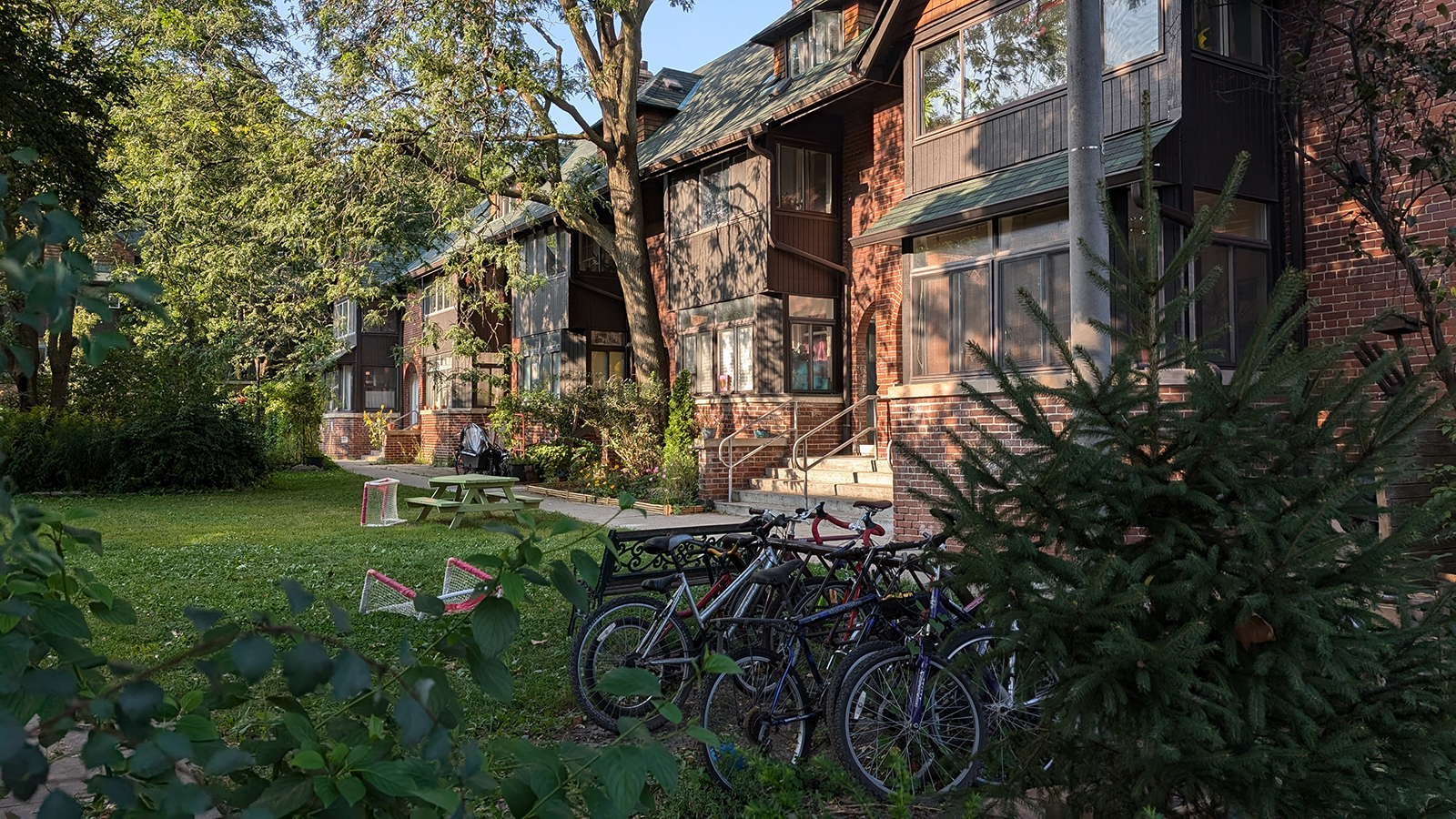 View of a row of houses in a green, shaded courtyard with large trees. The buildings have a mix of dark wood and brick facades with sloped roofs. Bicycles are parked in the foreground near a small spruce tree, and a picnic table and children’s mini hockey nets are set up on the lawn.