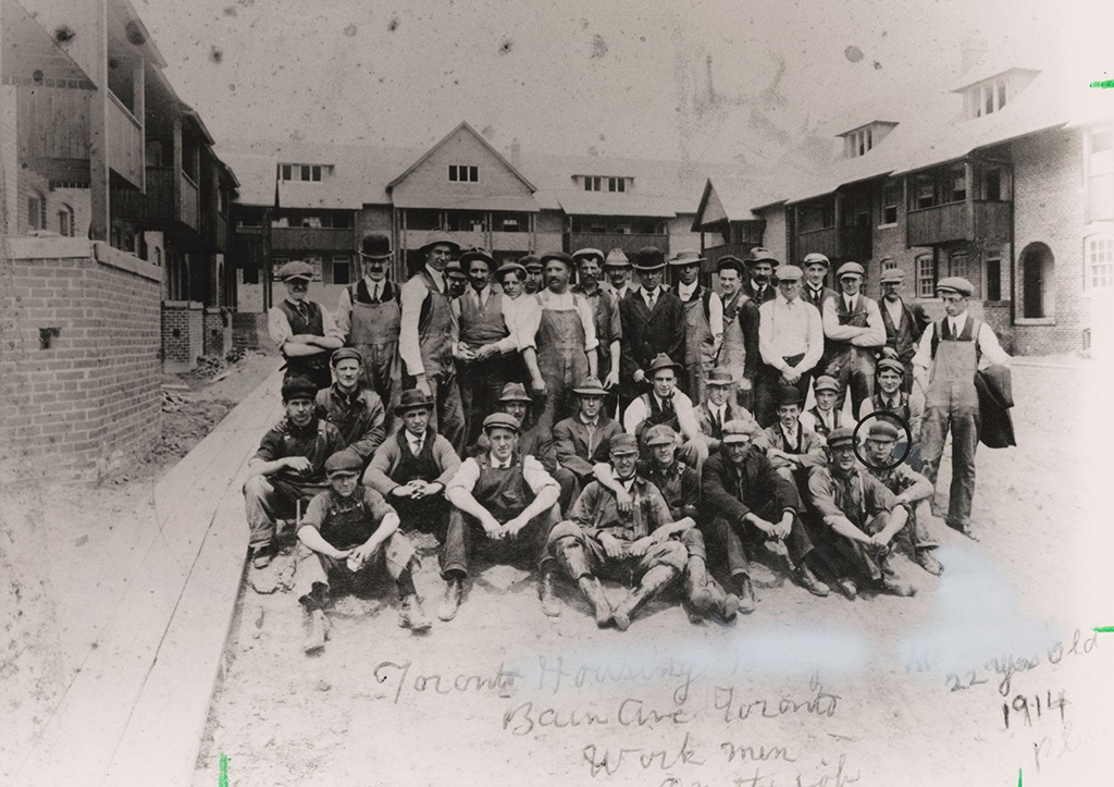 Historic black and white photograph from 1914 showing a group of construction workers posing in front of newly built housing units in Toronto. They are dressed in work attire including hats, vests, and overalls, stand and sit in rows, smiling at the camera. Handwritten text at the bottom of the photo reads ‘Toronto Housing Board, Bain Ave Toronto, Workmen 1914.