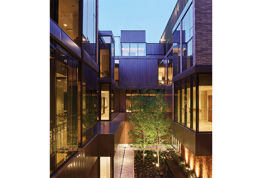 Evening view of a modern courtyard surrounded by a multi-story building with large glass windows. The courtyard features a small garden with trees and plants, illuminated by soft lighting.  Reflections from the glass surfaces highlight the architectural lines and stacked blocky massing.