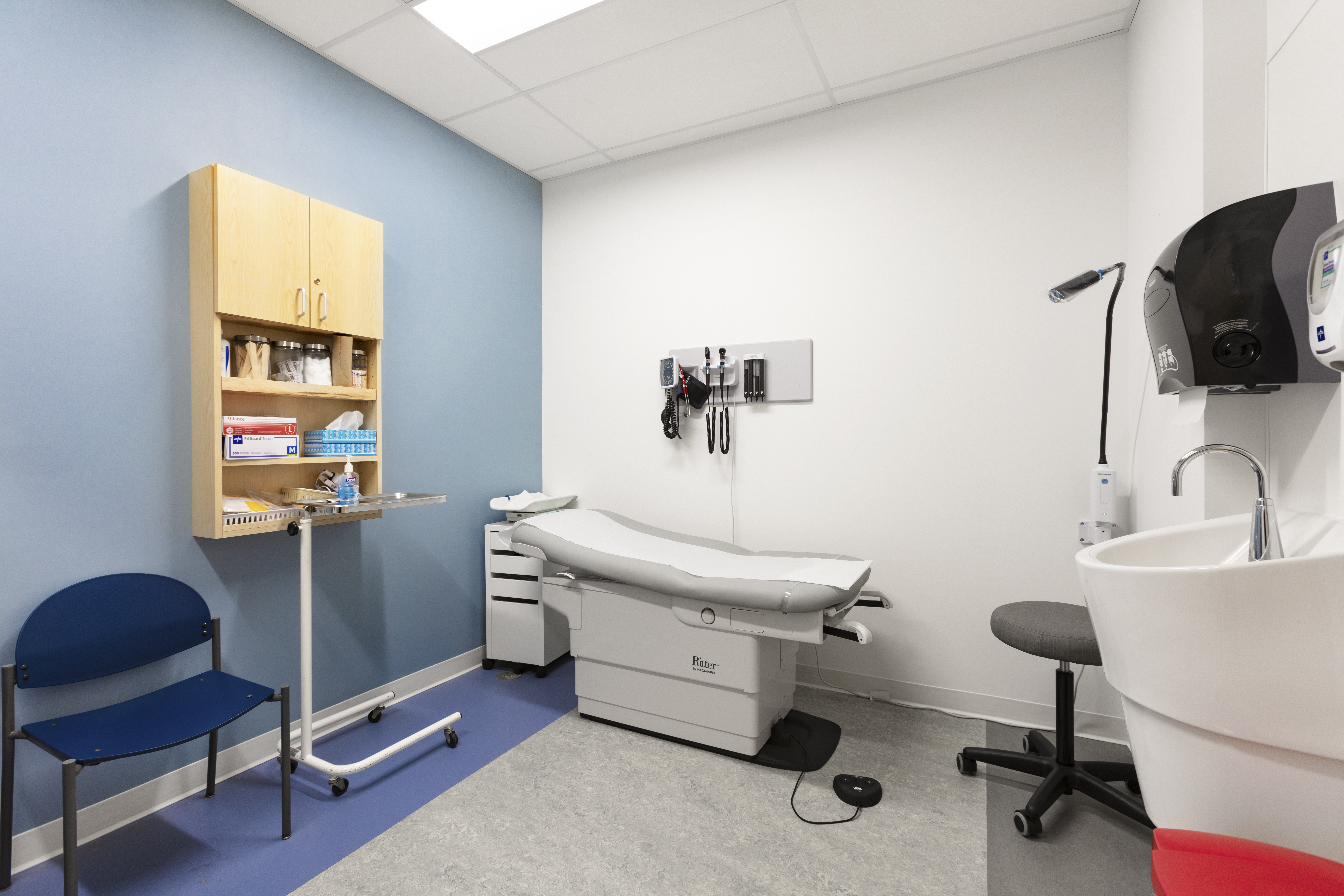 Interior of a medical examination room with a blue accent wall. The room contains a medical examination table, a mounted wall unit with medical tools, a small sink, and a blue chair. A storage cabinet with medical supplies is mounted on the wall, and a rolling tray table is positioned next to the examination bed  