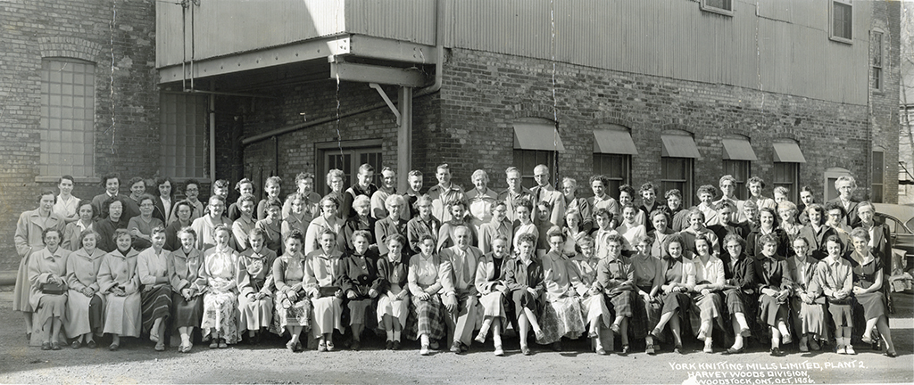 Black and white photograph of a large group of workers taken in October 1956. The group consists of men and women, mostly dressed in work attire, standing and sitting in rows outside a brick industrial building. The workers pose together for the camera, and writing in the bottom right corner of the photo provides details about the location and date.